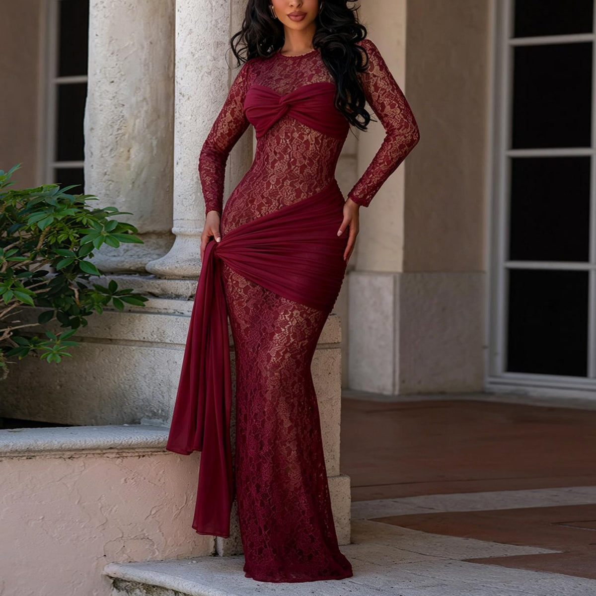 A woman models a long-sleeve burgundy lace evening gown with a sweetheart neckline and draped side detail, standing outdoors in front of a beige stone building.