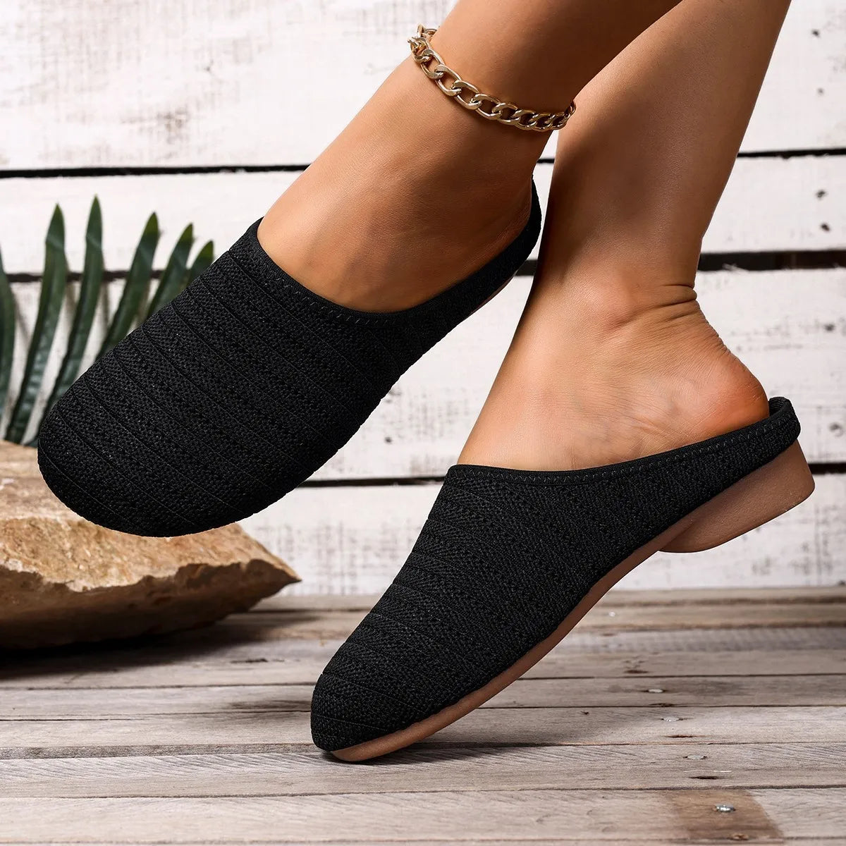 Black slip-on shoes worn by a person on a wooden floor with a plant in the background