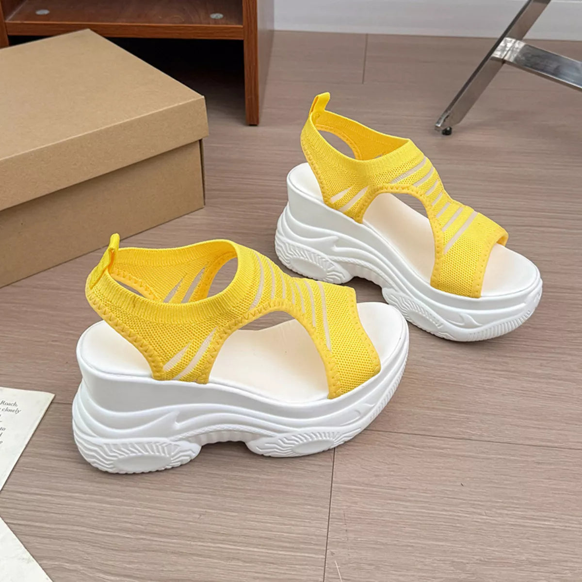 A pair of chunky platform sandals with bright yellow knit slingback straps and thick white platform soles, displayed on a light wood floor next to a cardboard shoe box.