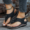 Black floral sandals worn with rolled-up jeans on a wooden surface.