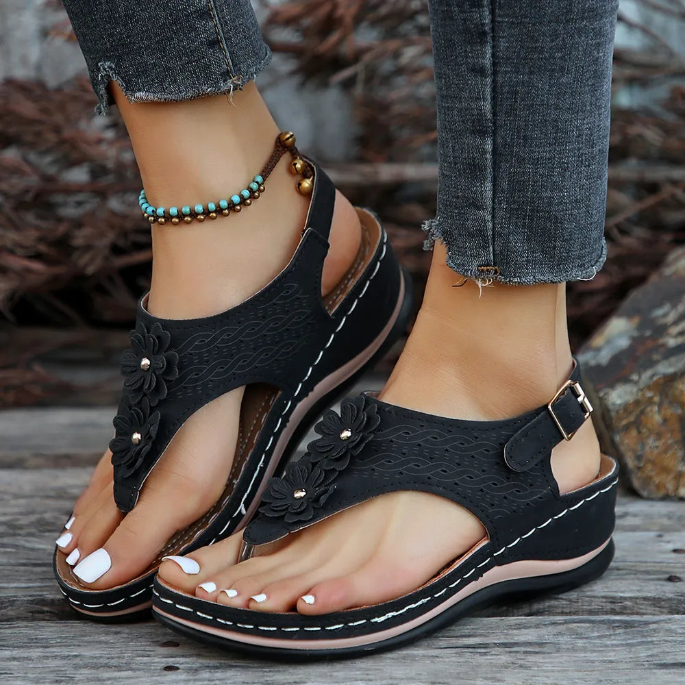 Black floral sandals worn with rolled-up jeans on a wooden surface.