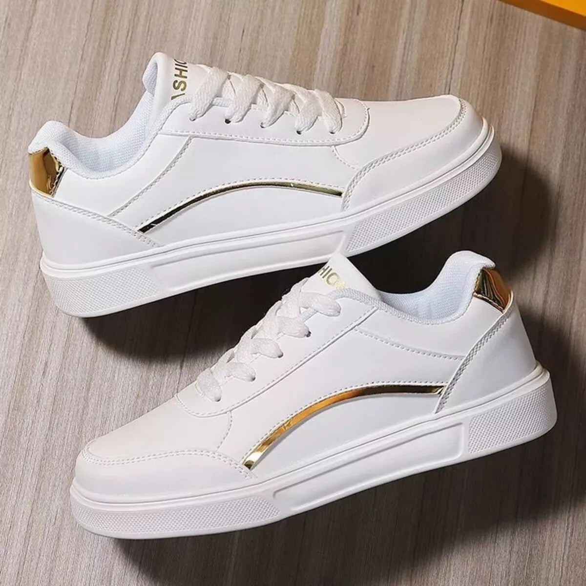 A pair of white low-top sneakers with metallic gold accents on the side and heel, displayed on a light wood floor-blackorvani.com