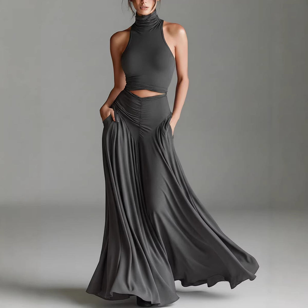 A woman modeling a dark gray two-piece outfit consisting of a sleeveless mock-neck crop top and a matching high-waisted, flowing maxi skirt with pockets, set against a neutral gray studio background.