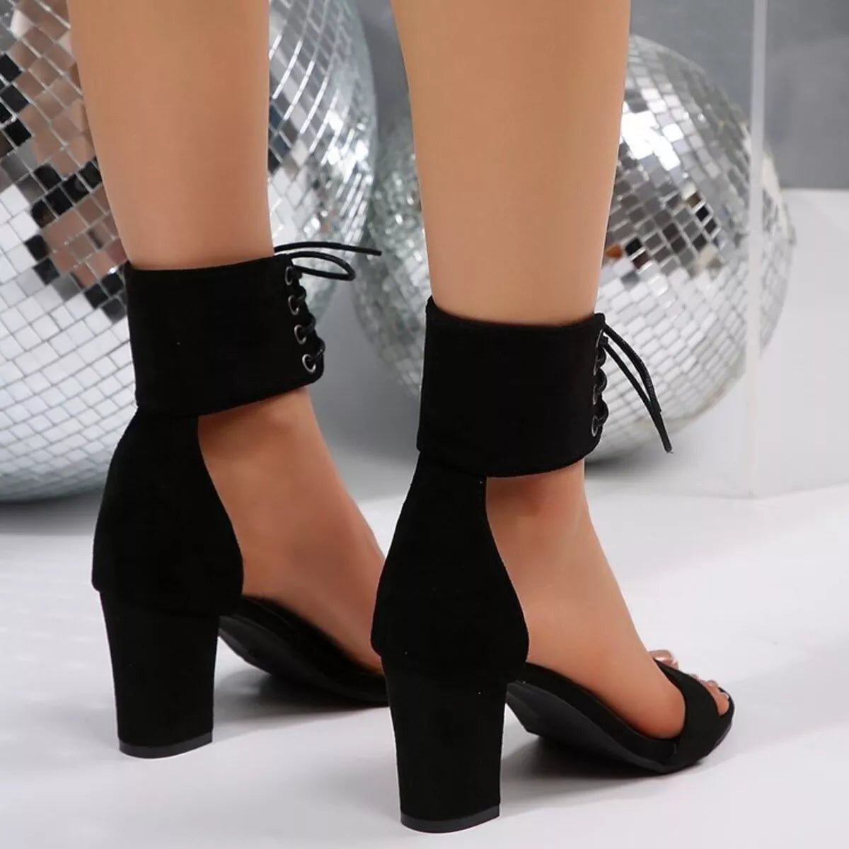 Black suede-look ankle-strap heeled sandals with lace-up front details, shown on a model's feet against a white floor with mirrored disco balls in the background-blackorvani.com