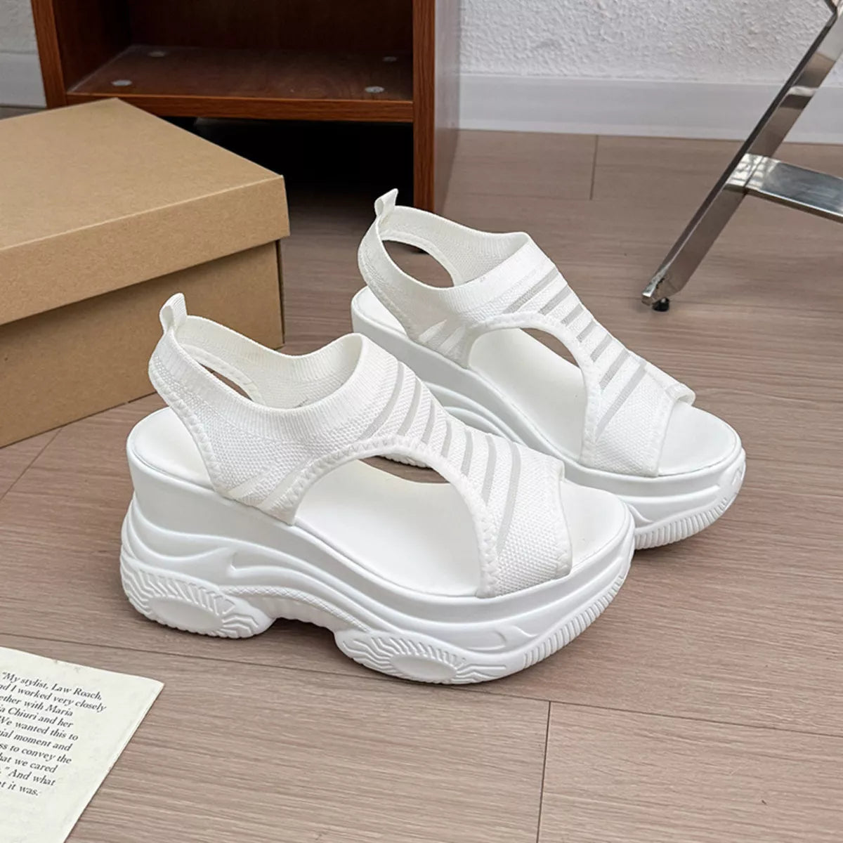 A pair of chunky white platform sandals with a textured knit slingback strap, displayed on a light wood floor next to a brown shoebox.