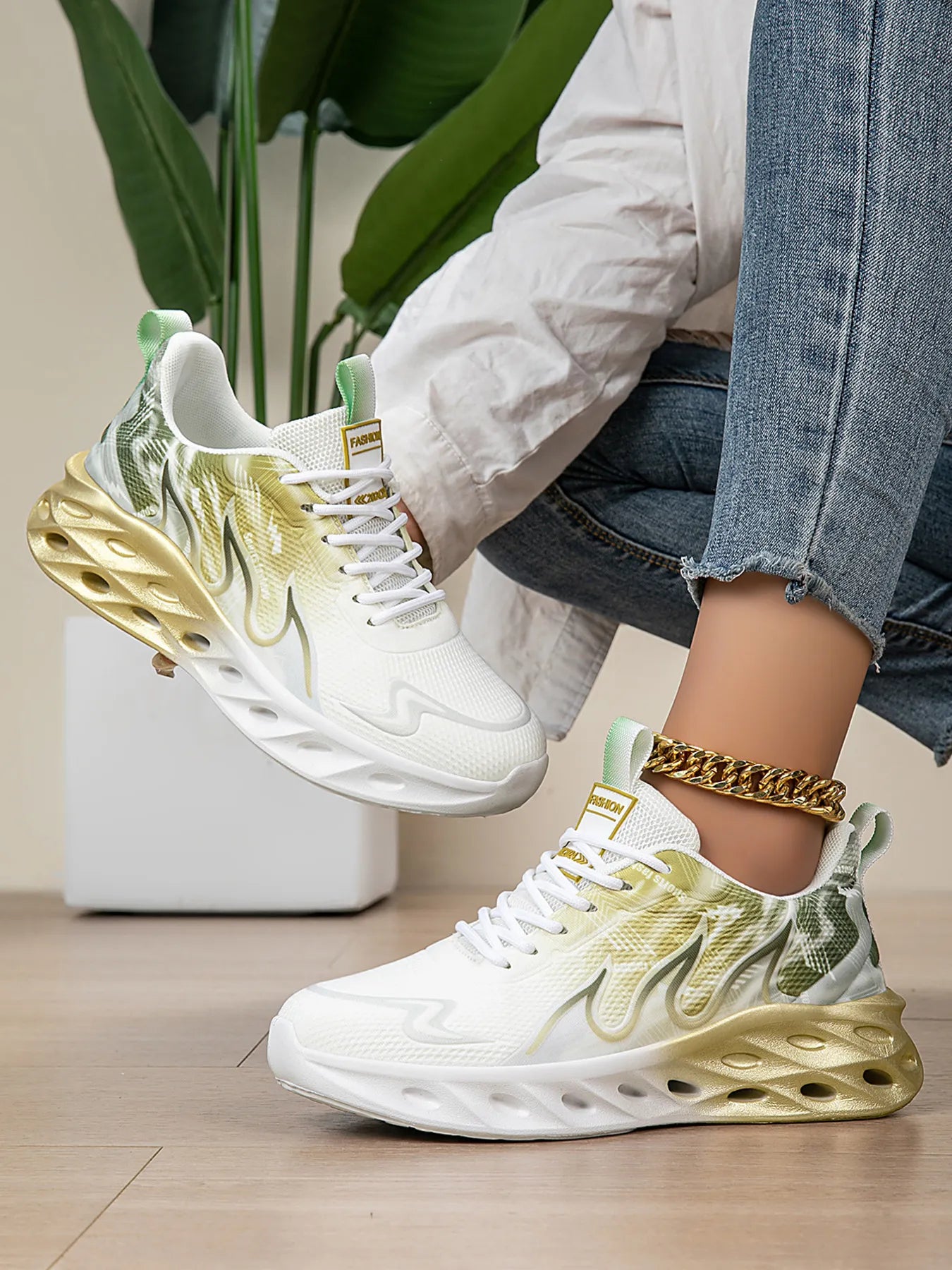 White and gold sneakers worn by a person sitting on a chair with a plant in the background.