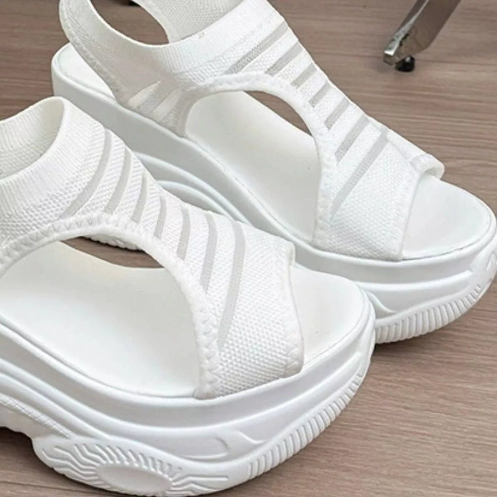 A pair of white open-toe platform sandals with wide woven straps and thick textured soles, photographed on a light wood floor.