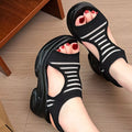 Black platform slide sandals with white horizontal stripes, shown on a person's feet on a light wood floor.
