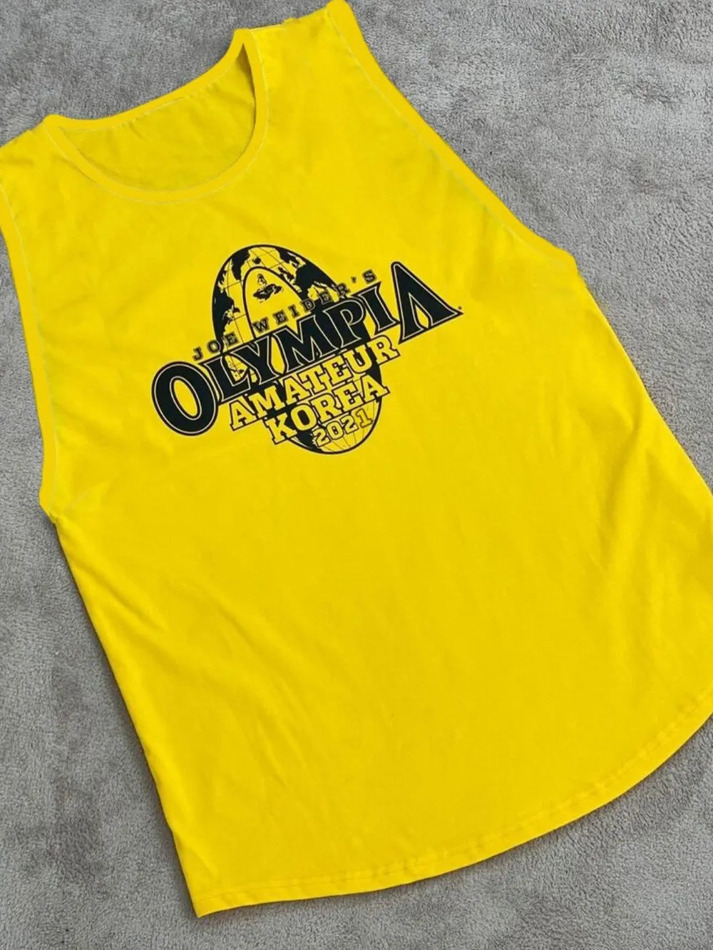 Yellow tank top with 'Olympia Amateur Korea 2023' print on a gray surface-blackorvani.com