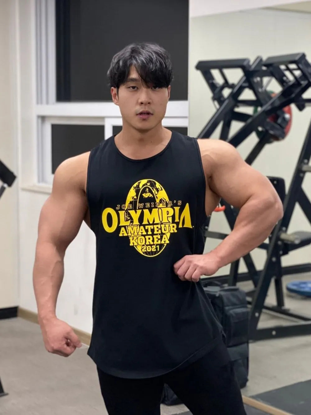 Muscular individual wearing a black tank top with 'Olympia Amateur Korea' text in a gym setting-blackorvani.com