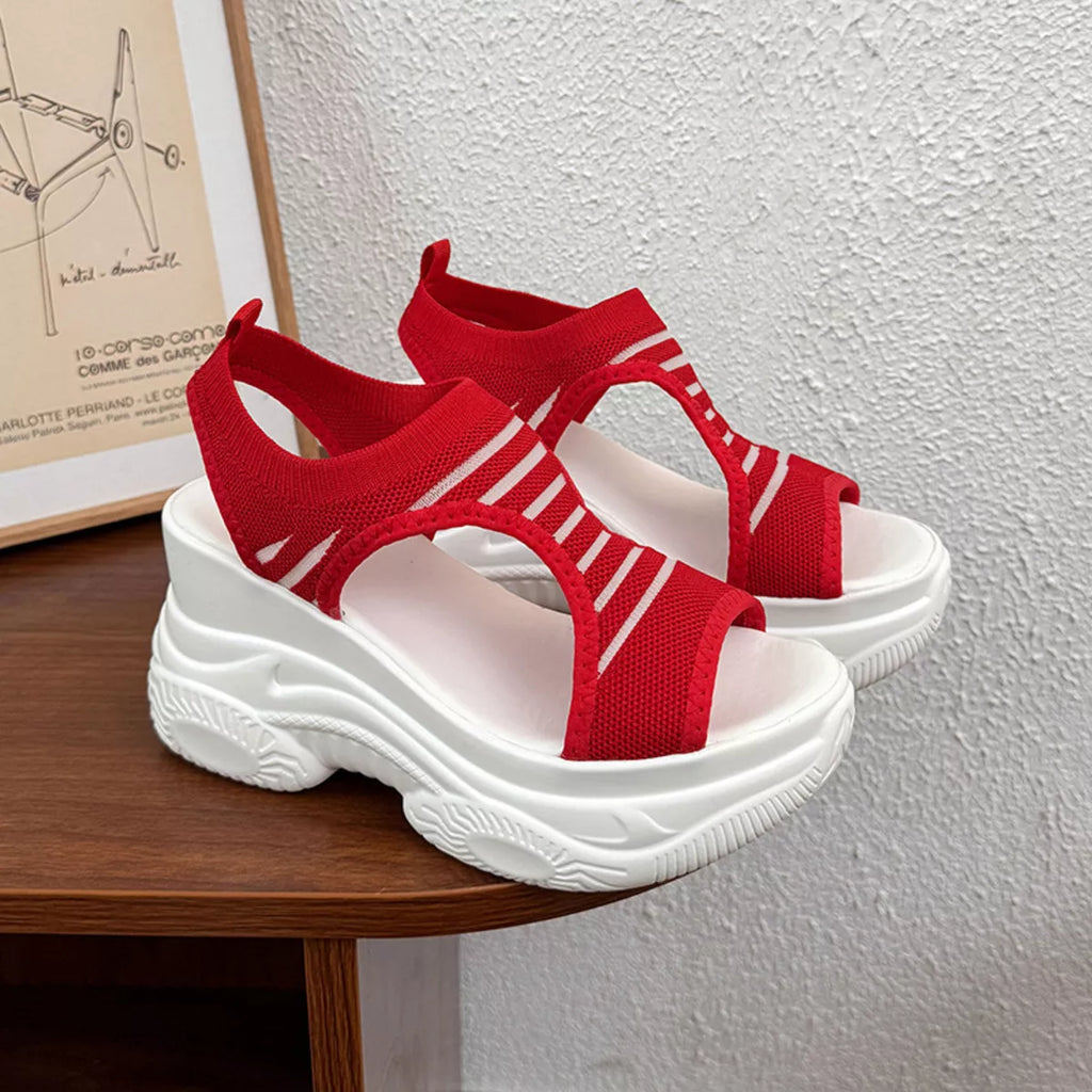 A pair of red knit slingback sandals with chunky white platform soles, displayed on a wooden table against a textured white wall.