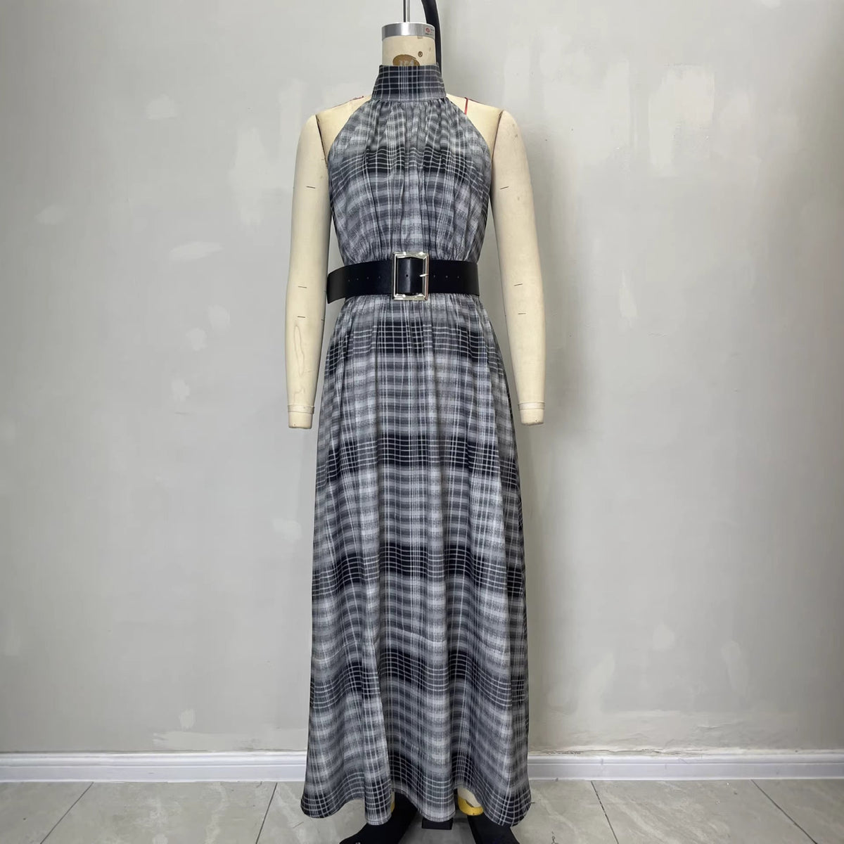 A sleeveless halter-neck maxi dress with a gray, black, and white plaid pattern, styled on a cream-colored mannequin with a black belt, set against a plain gray wall.