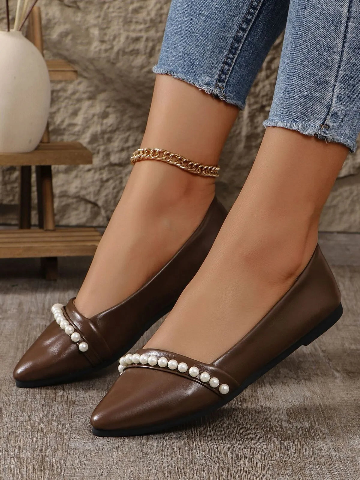 Brown pointed-toe flats with pearl embellishments worn by a person, with a blurred indoor background.
