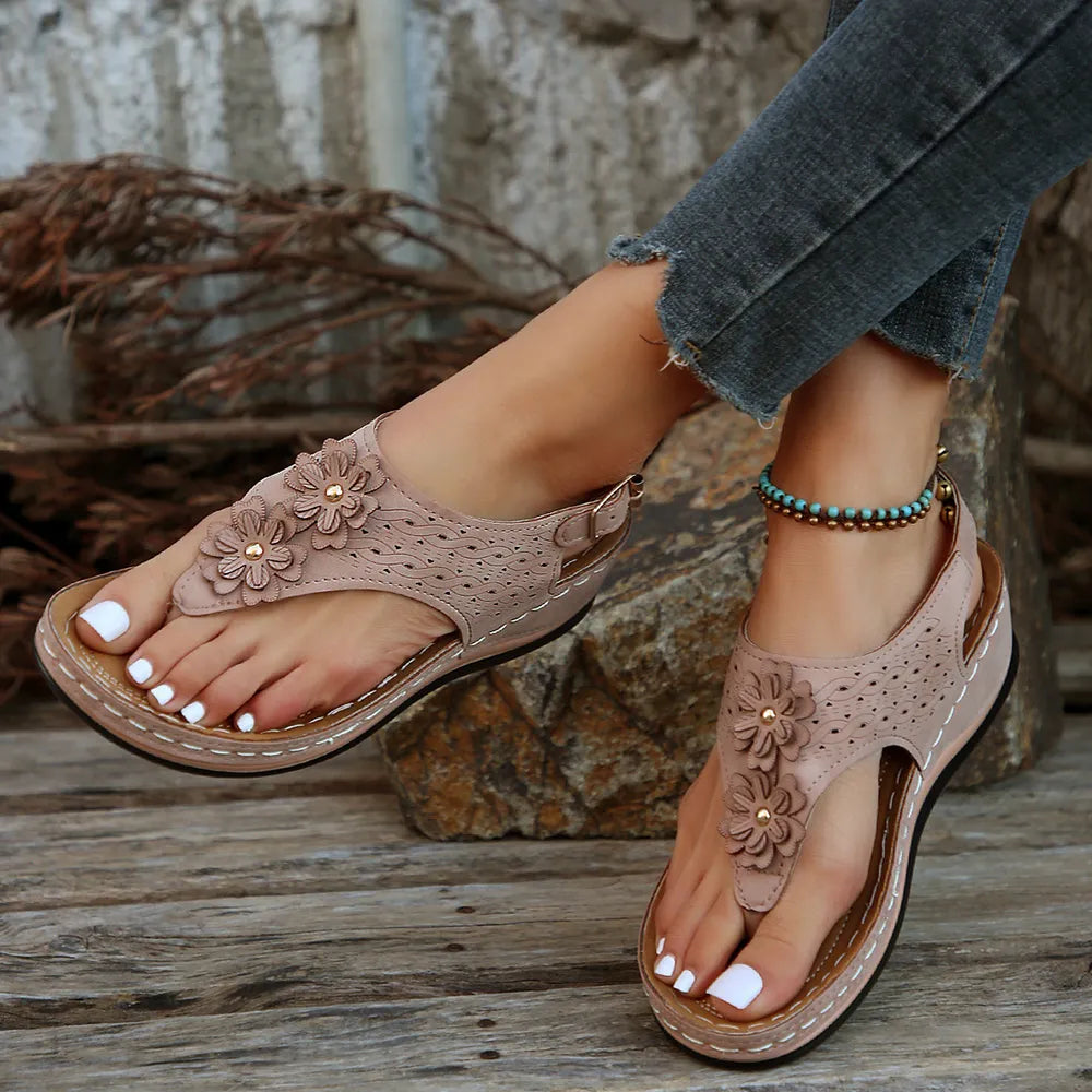 Beige sandals with floral designs worn by a person sitting on a stone surface.