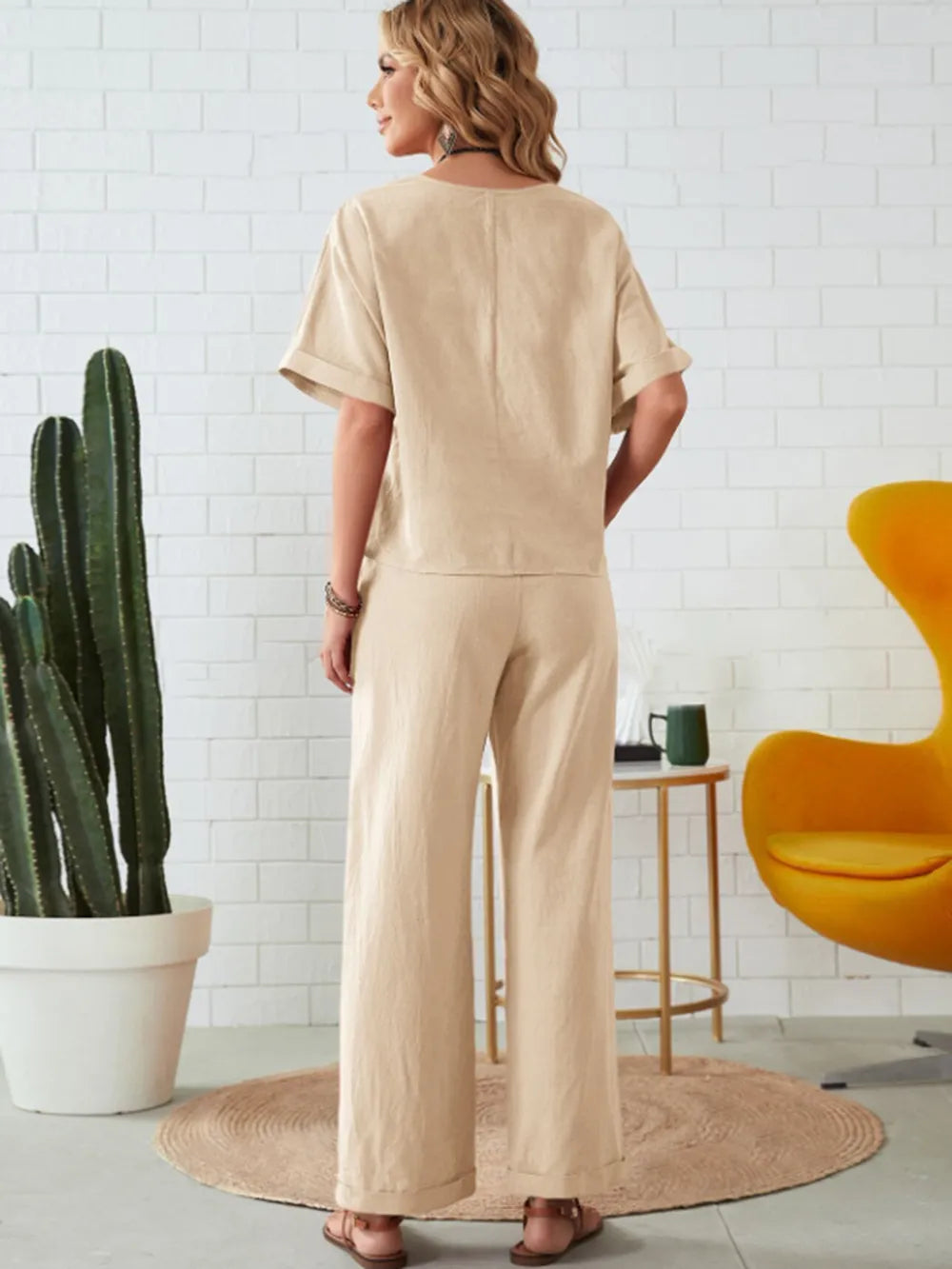 Woman wearing a beige jumpsuit standing in a room with a cactus plant and a yellow chair-blackorvani