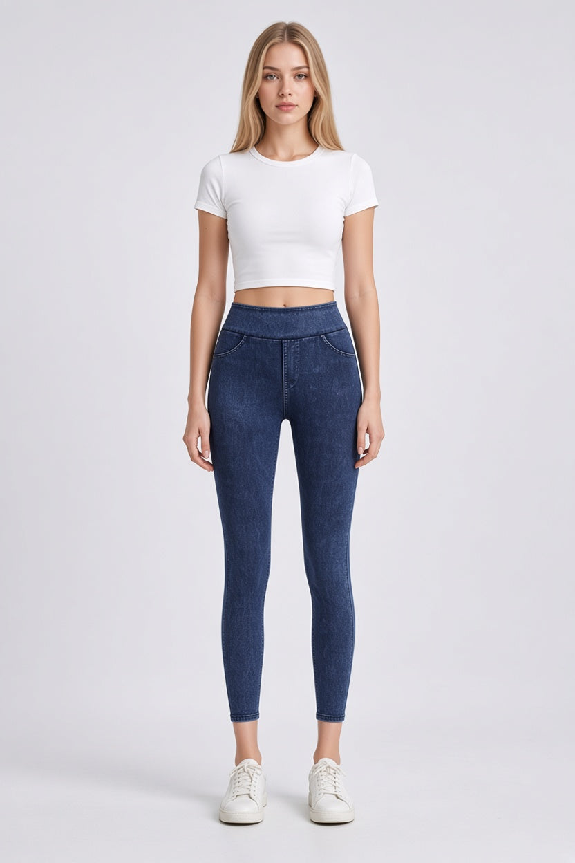A woman modeling a white short-sleeve crop top and dark blue high-waisted skinny jeans with white sneakers, standing against a light gray studio background.