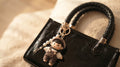 A close-up of a black leather handbag with a braided top handle and a dangling charm featuring a small doll wearing a white shirt and brown overalls, resting on a soft beige fabric background-blackorvani

