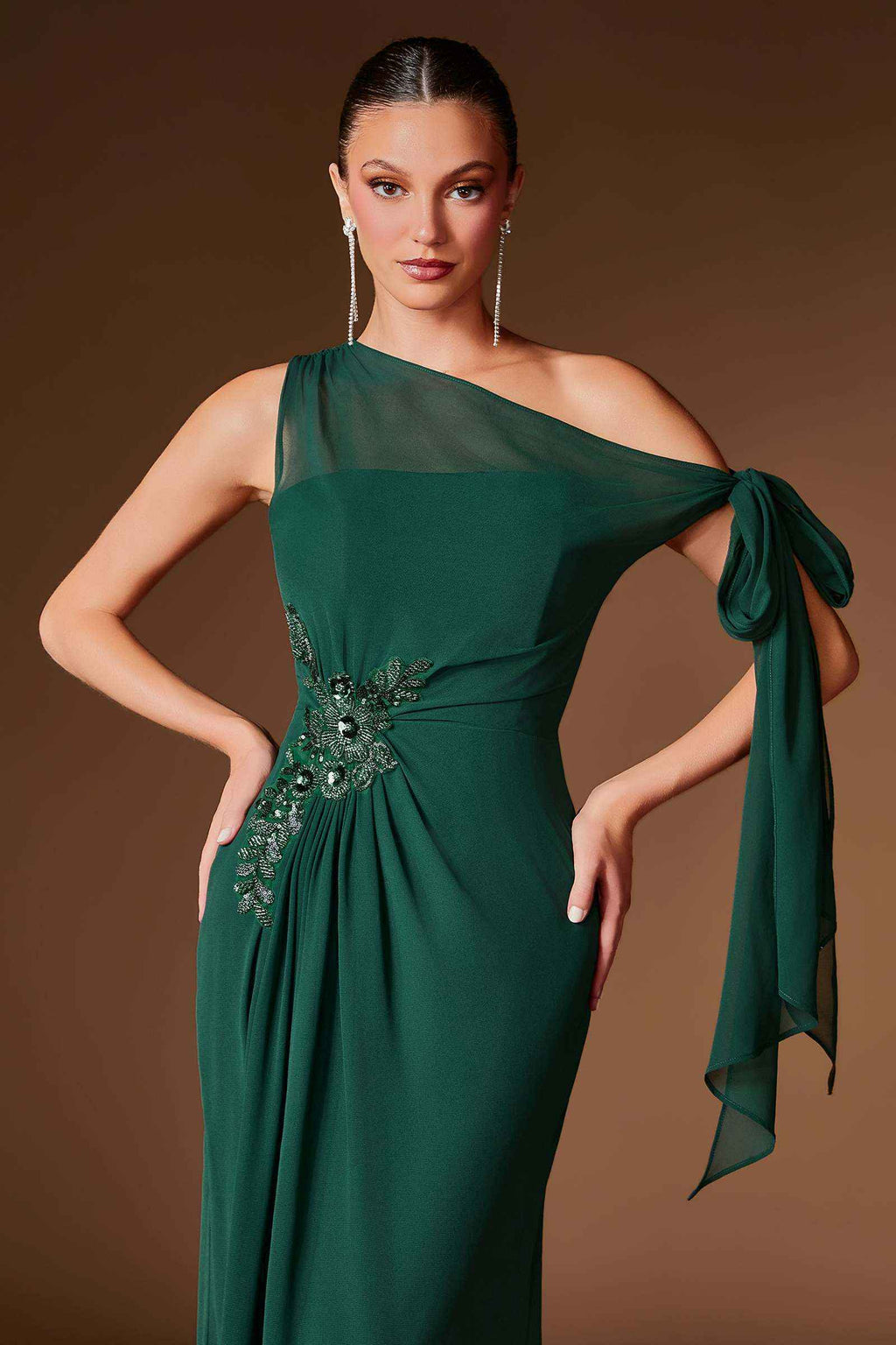 A woman modeling a dark green one-shoulder evening gown with a sheer mesh shoulder panel, a side tie detail, and an embellished waistband, set against a solid brown studio background-blackorvani