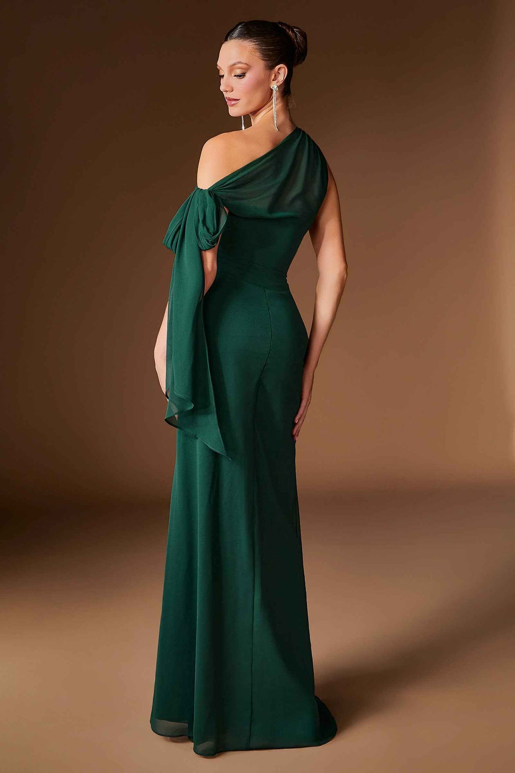 A woman modeling a floor-length emerald green one-shoulder gown with a draped bow detail on the bodice, photographed against a warm brown studio backdrop-blackorvani