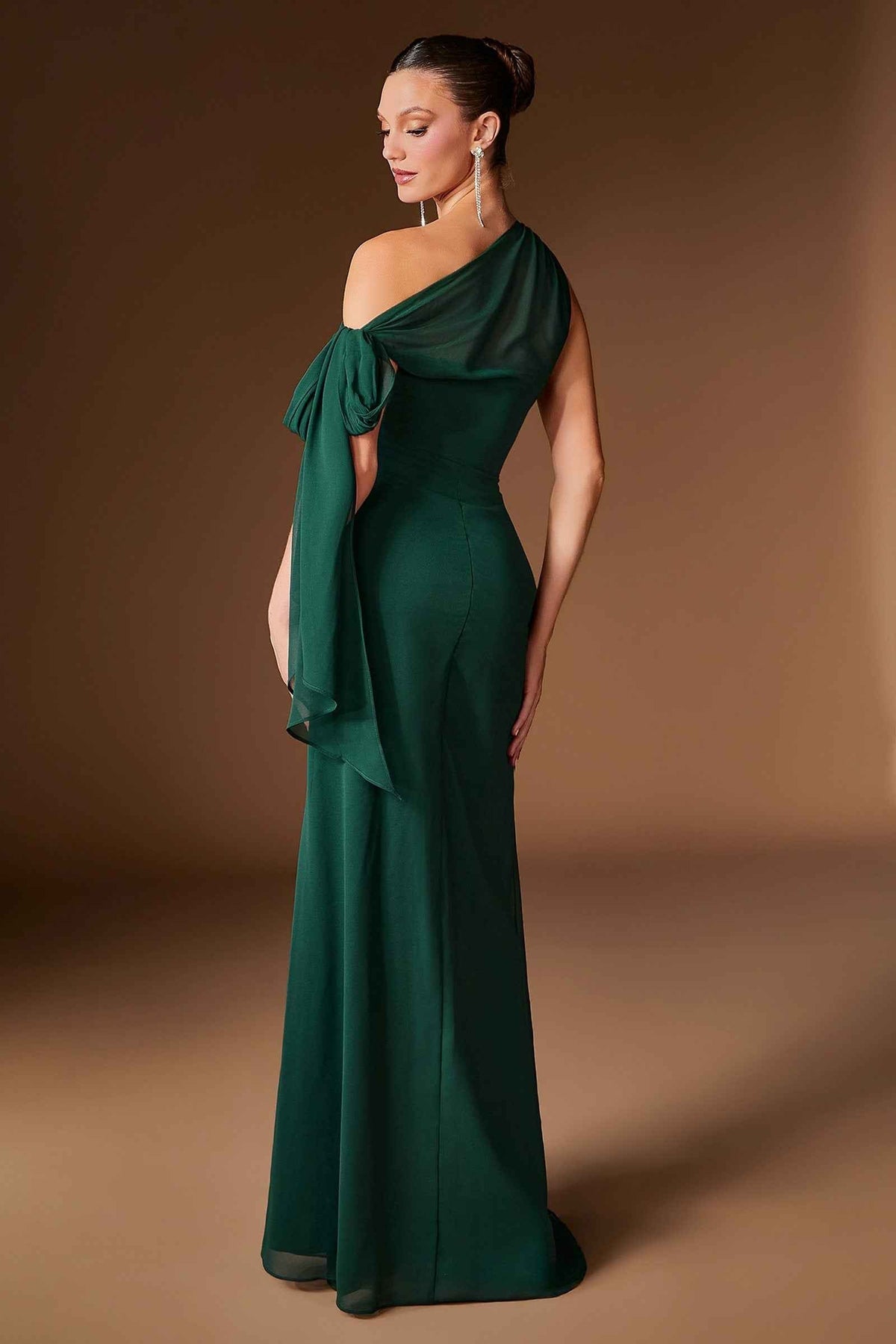 A woman modeling a floor-length emerald green one-shoulder gown with a draped bow detail on the bodice, photographed against a warm brown studio backdrop-blackorvani