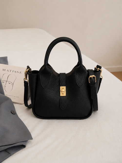 A black textured leather handbag with gold-tone hardware, featuring a top handle, a front strap closure with a gold clasp, and a detachable shoulder strap, styled on a white bed next to a gray garment and a book-BlackOrvani