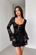 A woman modeling a black sheer floral lace mini dress with long flared sleeves, a scoop neckline, and a front keyhole cutout with ties-BlAcKORvAni