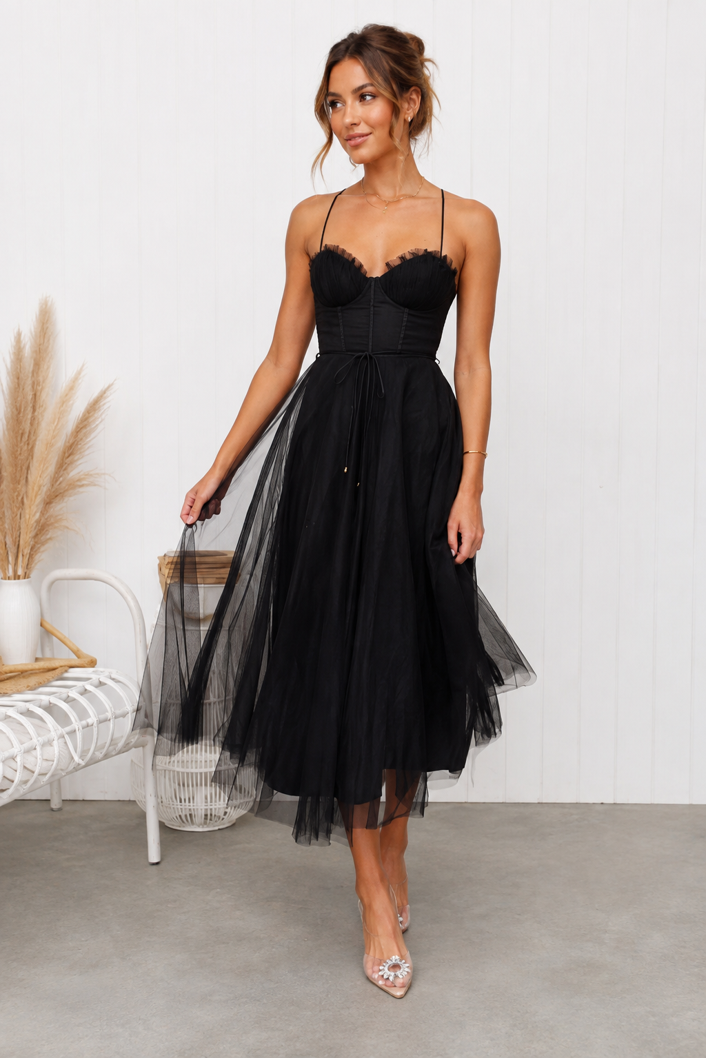 A woman models a black spaghetti-strap midi dress with a lace-trimmed sweetheart bodice, corset-style waist, and a layered tulle skirt, standing in a bright room with a white paneled wall and concrete floor-blackorvani.com