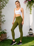 A woman stands outdoors on grass in front of a white wall, wearing olive green high-waisted athletic leggings with mesh side panels and a white sports bra.-blackorvani
