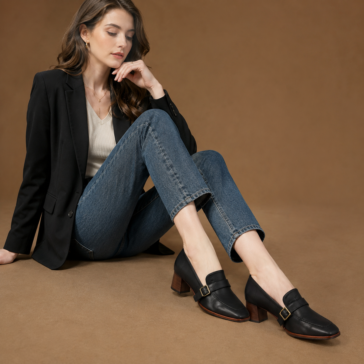 A woman sits on a brown studio floor wearing black leather loafers with a buckle strap, blue jeans, and a black blazer-blackorvani