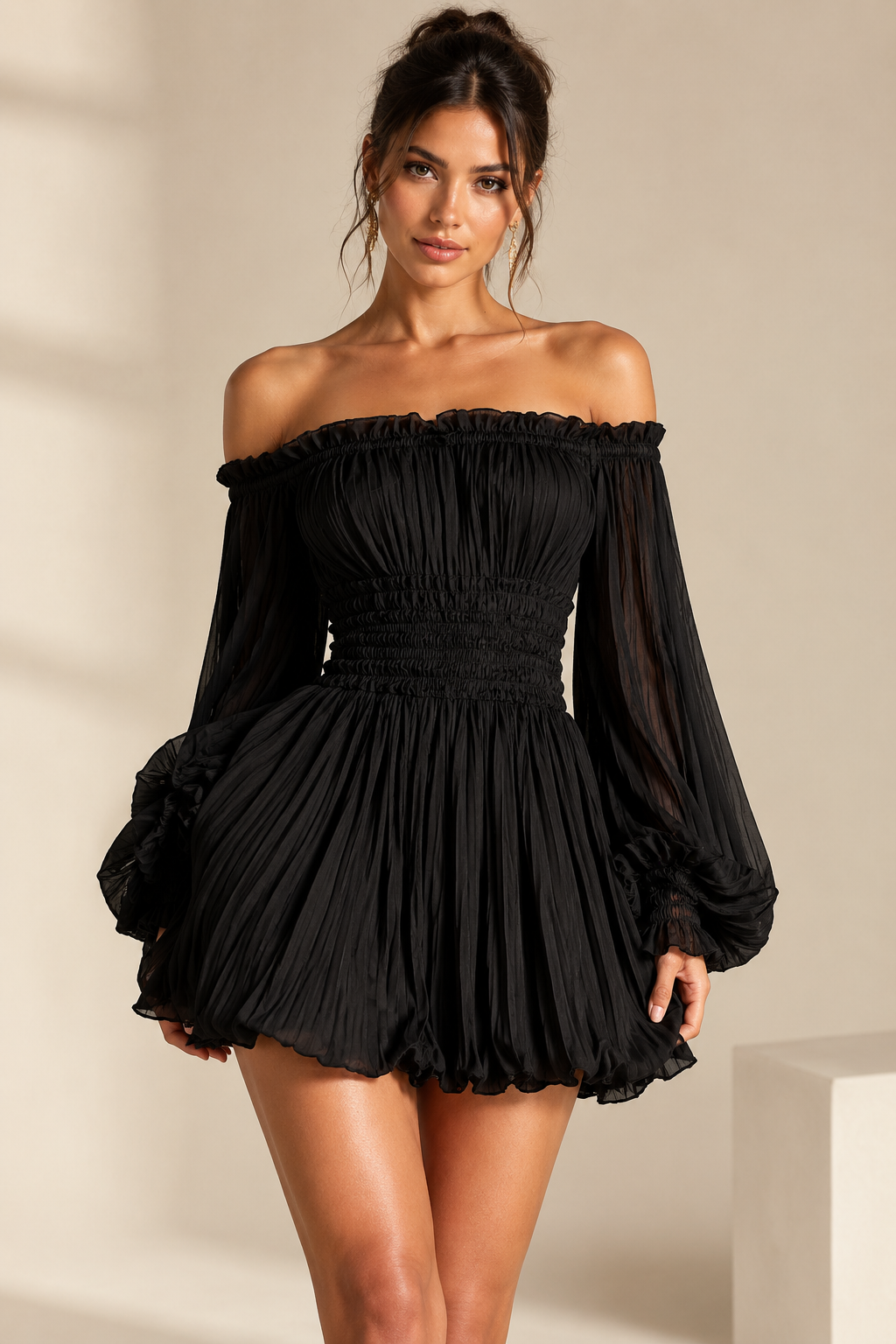 A woman modeling a black off-the-shoulder mini dress with long sheer puff sleeves, a smocked bodice, and a pleated flared skirt against a warm beige studio background.