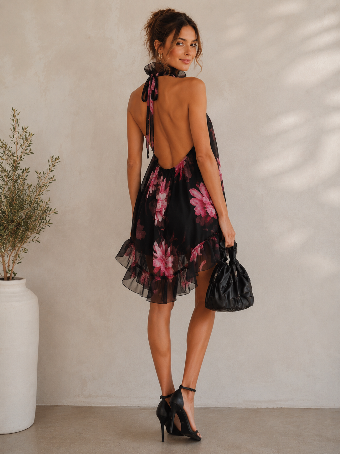 A woman stands with her back to the camera wearing a black halter mini dress with a pink floral print and an open back. She is holding a black handbag and wearing black high heels, standing in front of a textured beige wall with a potted plant on the left.