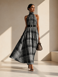 A model walks in a minimalist beige studio wearing a sleeveless halter-neck maxi dress with a black and gray plaid pattern, a black belt with a silver buckle, and holding a brown handbag.