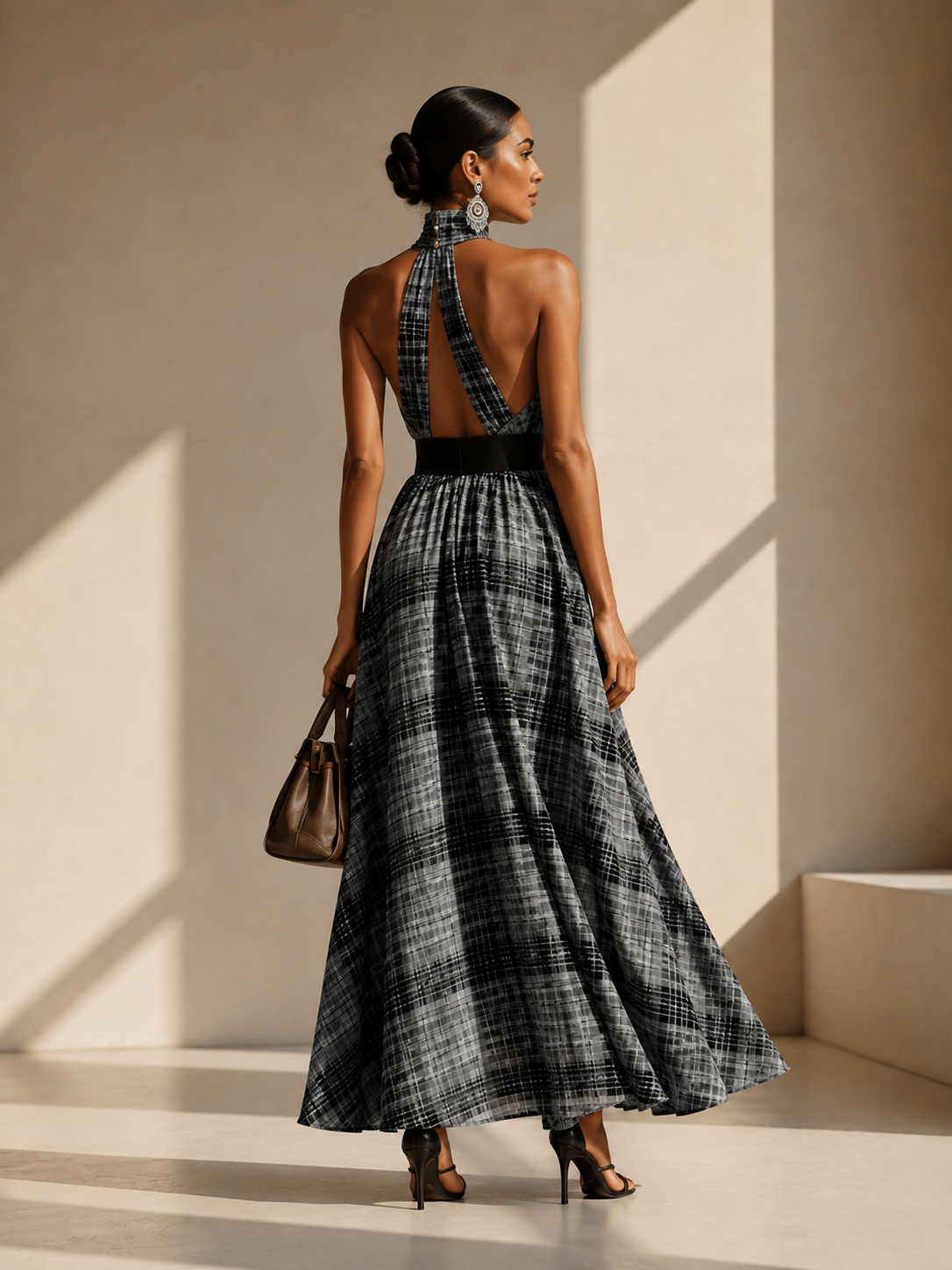 A woman stands with her back to the camera wearing a long halter-neck plaid dress in black, gray, and white, holding a brown handbag in a minimalist beige room with dramatic shadows.