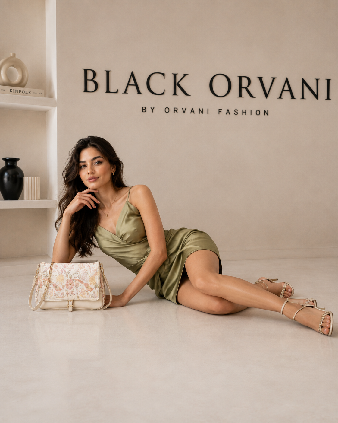 A woman in an olive green satin dress reclines on a light beige floor next to a cream floral-patterned handbag,BLACKOrVaNi