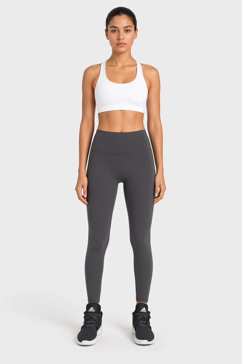 A woman stands against a light gray studio background wearing a white sports bra, dark gray high-waisted leggings, and black athletic sneakers.