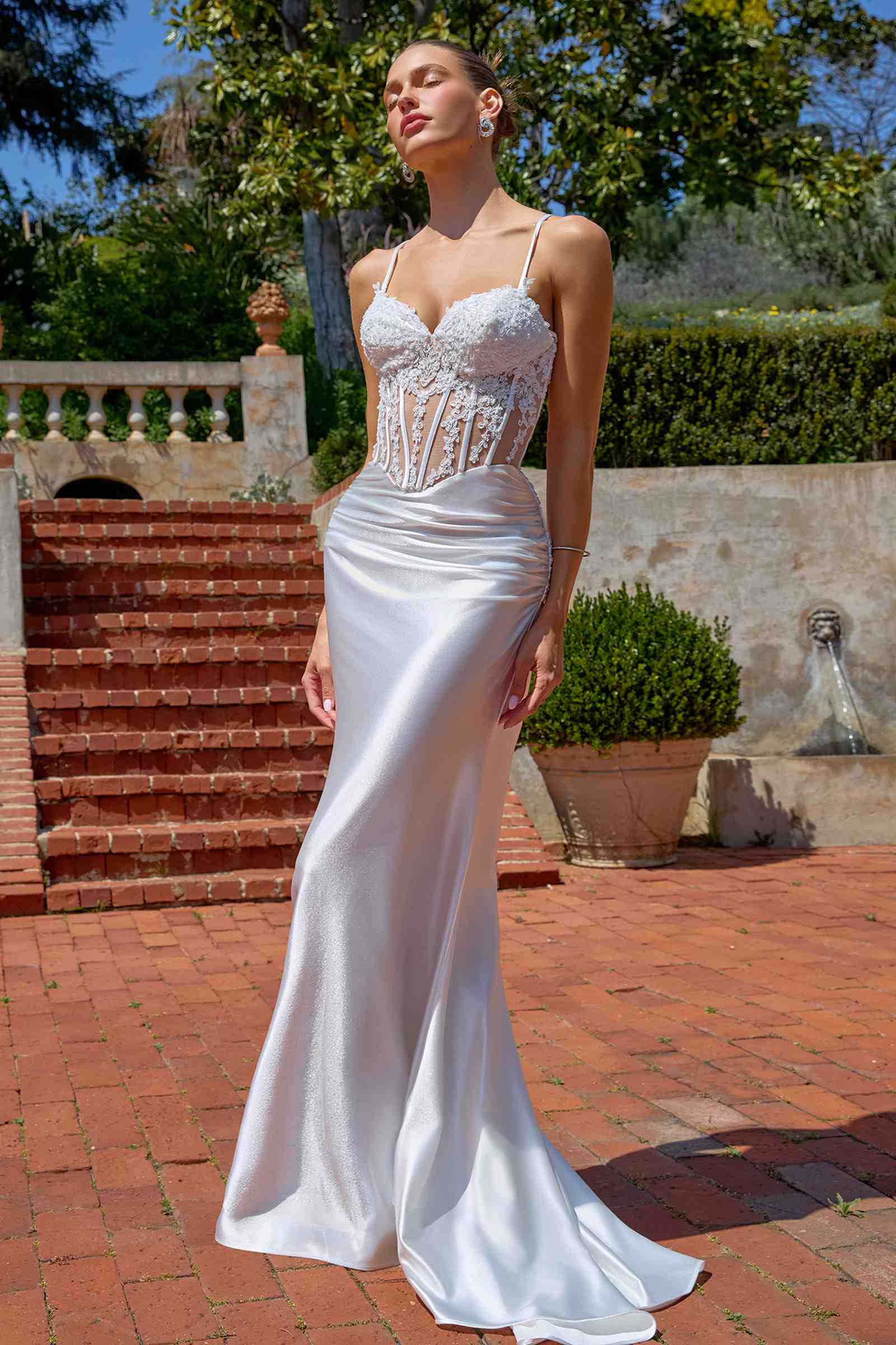 Woman in a white wedding dress standing outdoors with greenery and a fountain in the background.