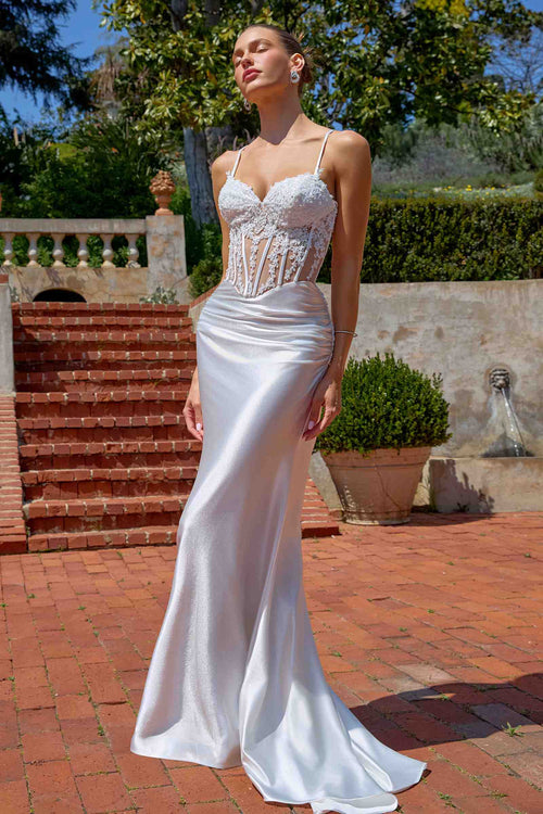 Woman in a white wedding dress standing outdoors with greenery and a fountain in the background.