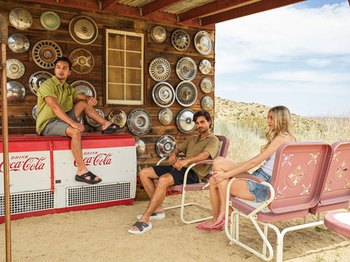 Three people sitting in a desert setting with a vintage Coca-Cola cooler and wall of hubcaps-blackorvani