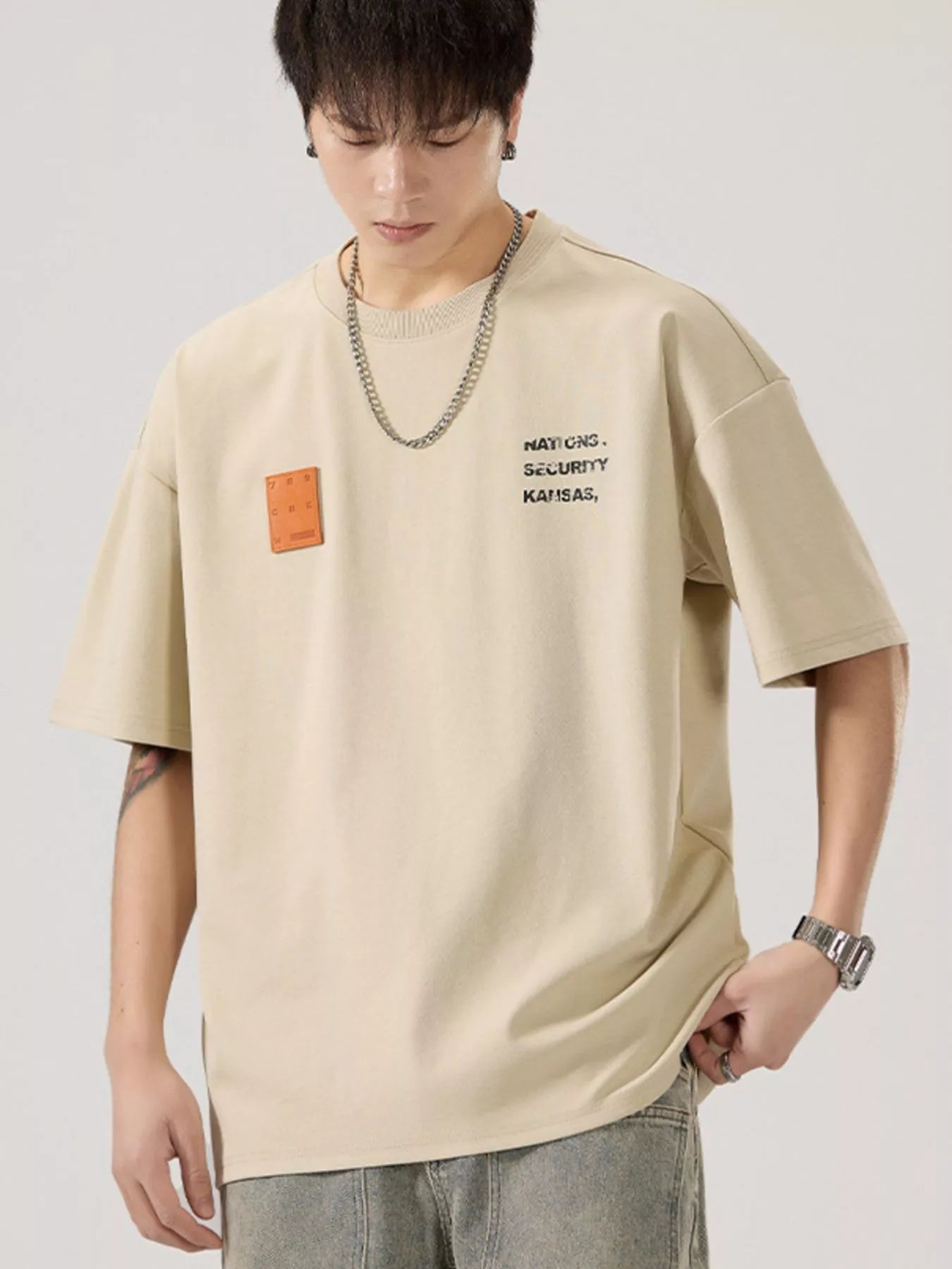 men  wearing a beige t-shirt with text and a patch on a plain background