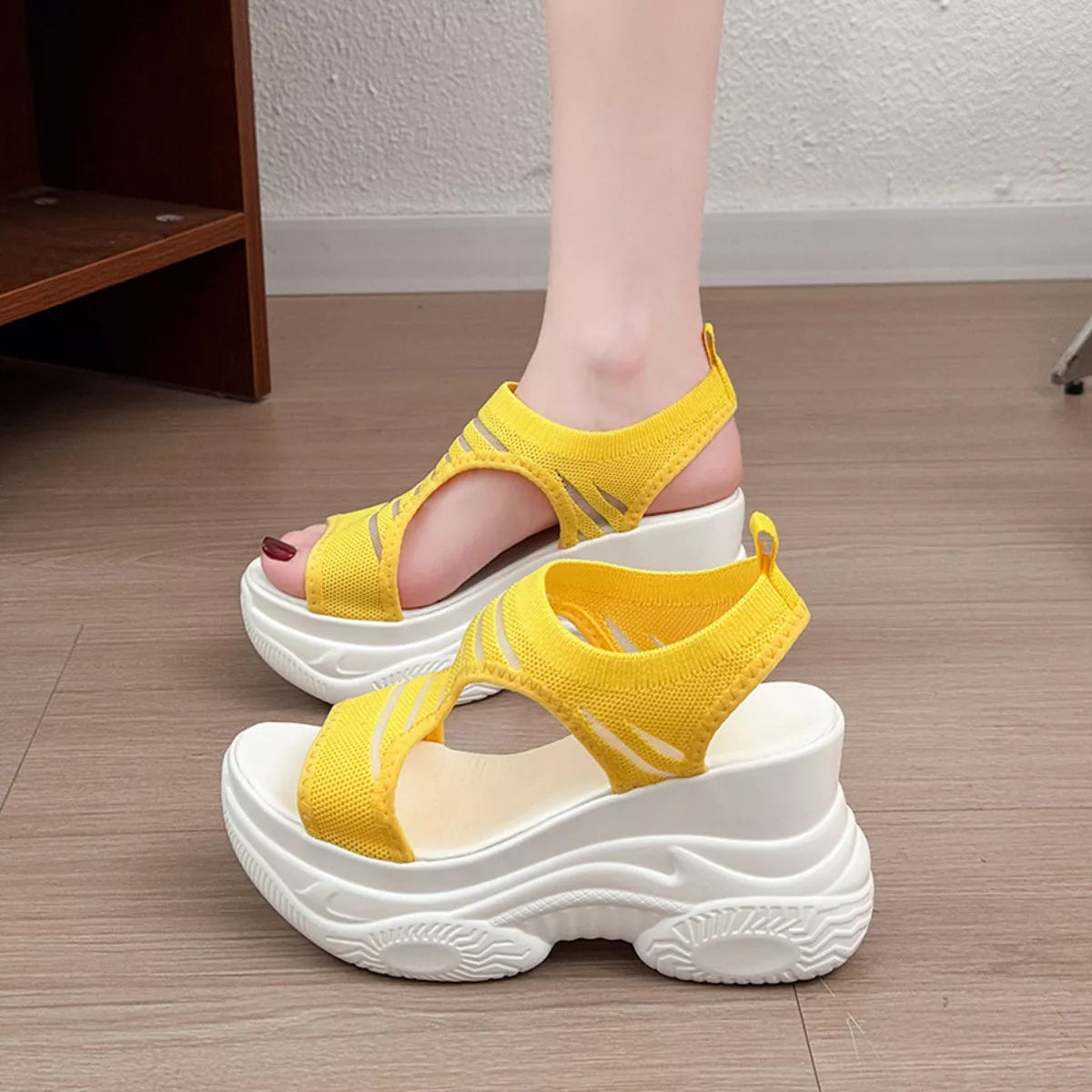 A pair of women's platform sandals in bright yellow and white, shown with one being worn on a foot and the other placed on a wooden floor.