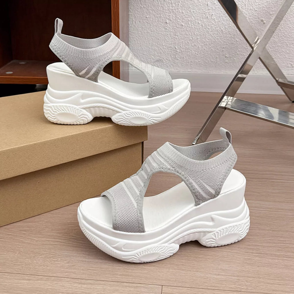 A pair of chunky platform sandals with white soles and light gray knit uppers, displayed on a wooden floor next to a cardboard shoebox.