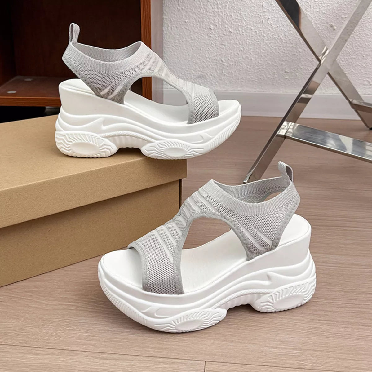 A pair of chunky platform sandals with white soles and light gray knit uppers, displayed on a wooden floor next to a cardboard shoebox.