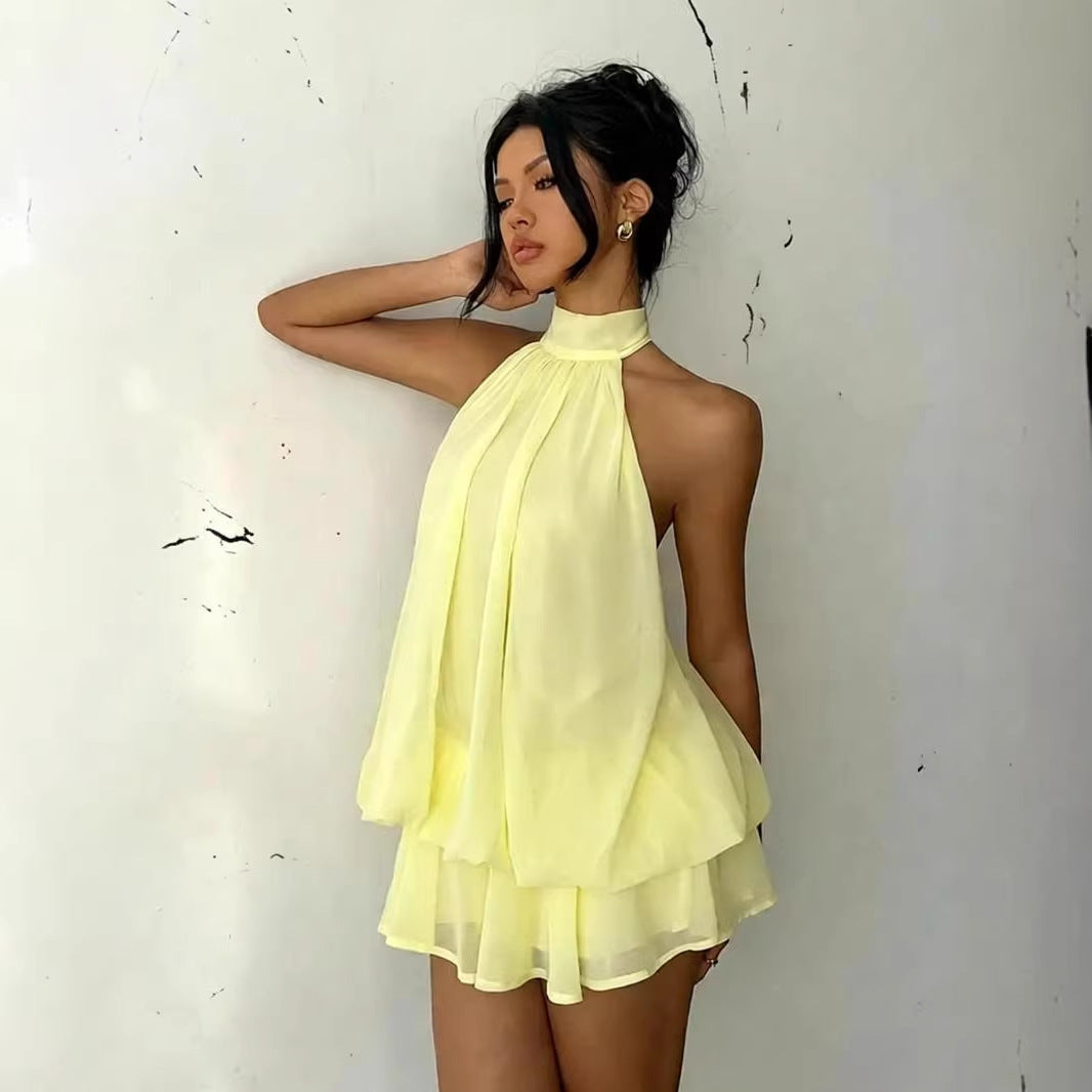A woman wearing a pale yellow halter-neck mini dress with a layered ruffle skirt, posing against a white wall.