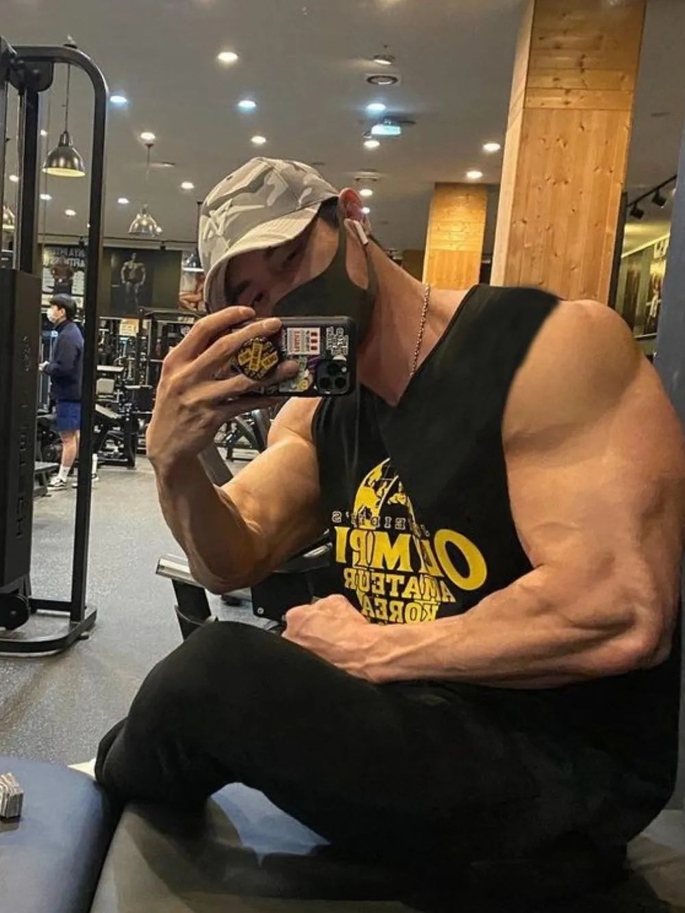 Person taking a mirror selfie in a gym wearing a black tank top with yellow text-blackorvani.com