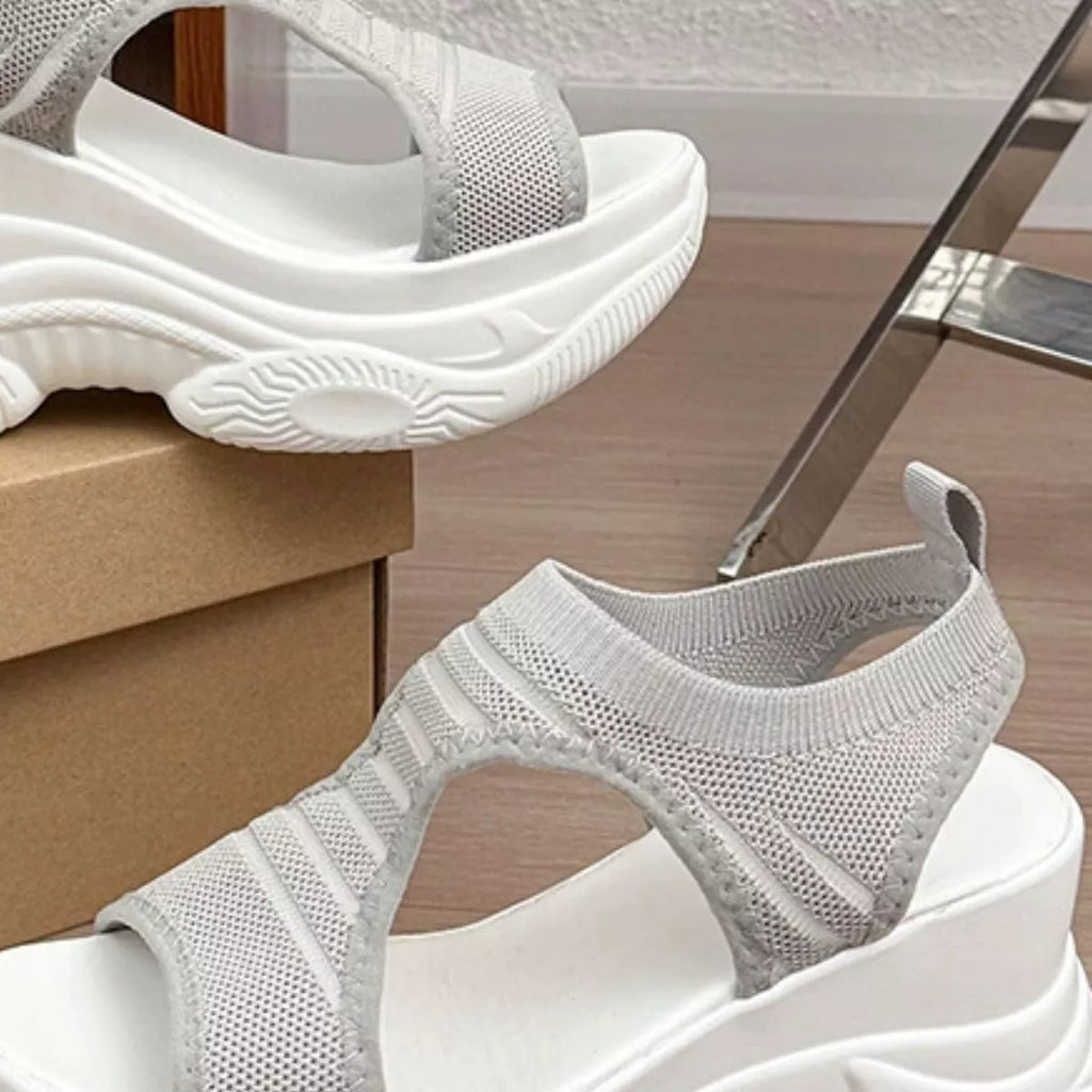 A pair of chunky platform sandals with light gray knit upper straps and white soles, shown on a wooden floor with one sandal resting on a cardboard box.