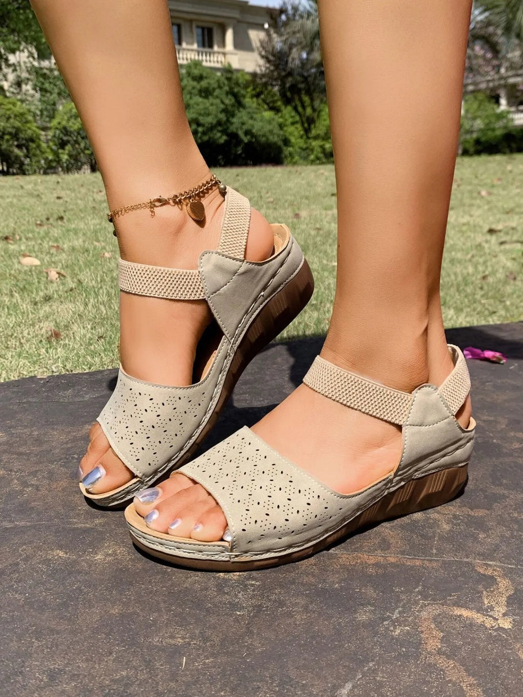Beige perforated wedge sandals with mesh ankle straps, shown on a model's feet outdoors on a dark stone surface with grass in the background.