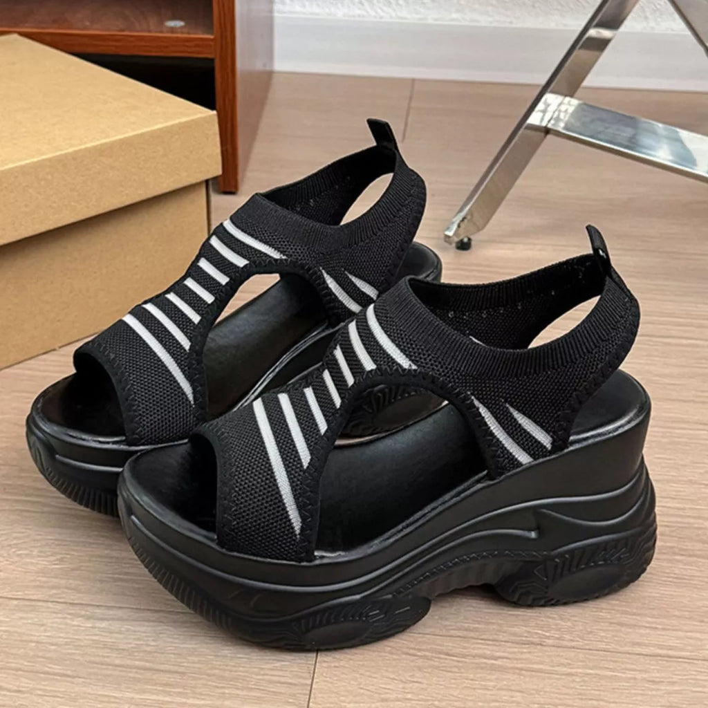 A pair of black platform wedge sandals with white diagonal stripe details on the straps, shown on a light wood floor next to a shoebox.