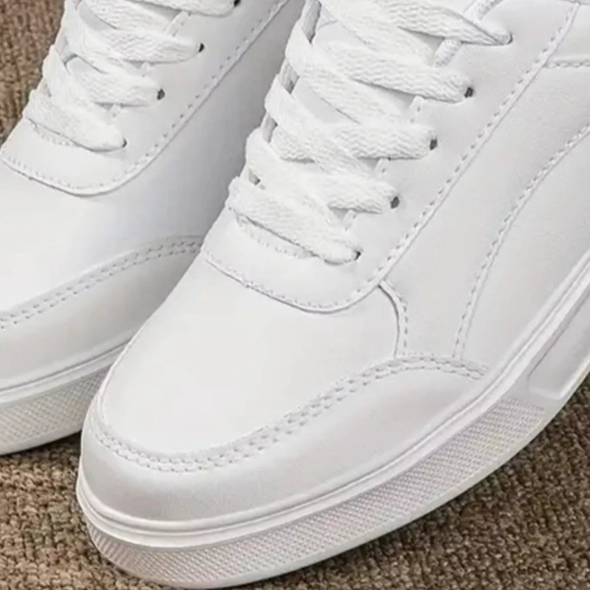 A close-up of a pair of white low-top sneakers with matching white laces, showing the toe cap, side panel, and textured sole on a neutral carpet background-blackorvani.com