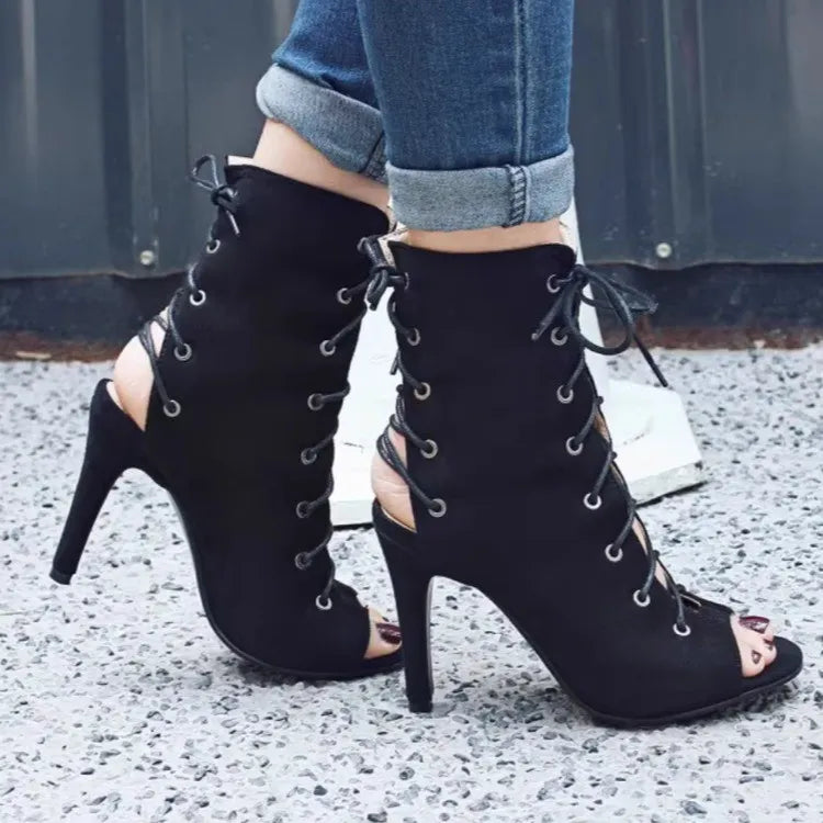Black lace-up high-heeled shoes worn with rolled-up jeans on a textured surface.