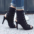 Black lace-up high-heeled shoes worn with rolled-up jeans on a textured surface.